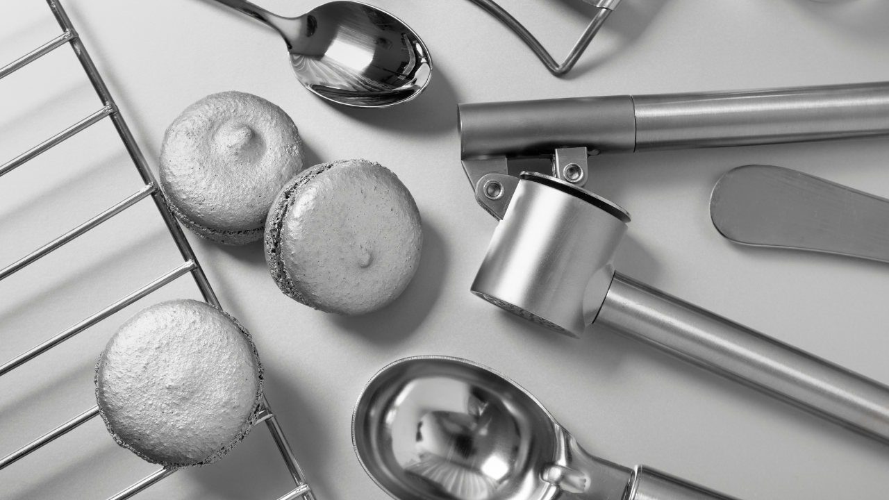 Utensílios gastronômicos em aço inox dispostos sobre superfície clara, acompanhados de macarons, em composição minimalista.