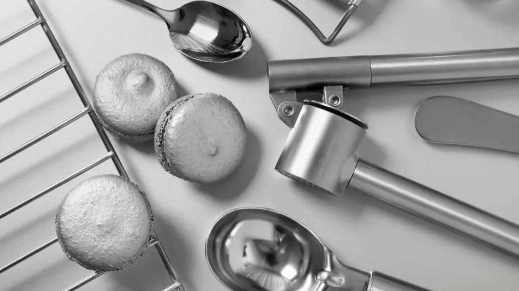 Utensílios de cozinha em aço inox sobre bancada, incluindo colheres metálicas, pegador de sorvete e grade, ao lado de doces prateados.