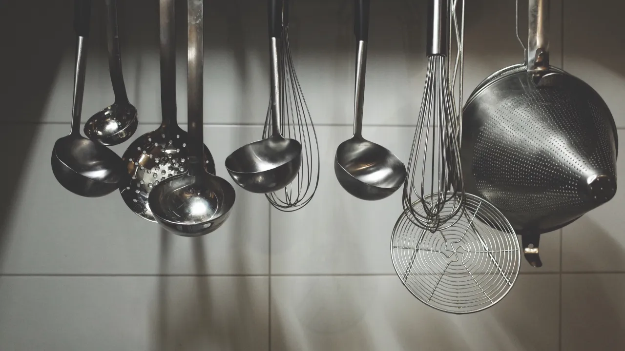 Utensílios gastronômicos em aço inox pendurados na parede, como conchas, escumadeiras e batedores, organizados em cozinha profissional.
